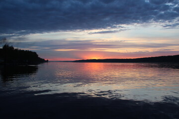 Obraz premium Tranquil Sunset Over Calm Water with Vibrant Orange and Purple Sky Reflections and Dock Extending Toward Tree-Lined Horizon