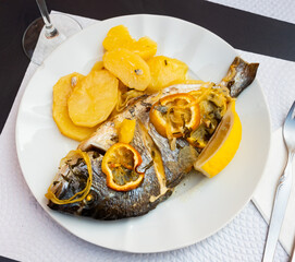 Carcass whole roasted dorado fish with baked potato is served on plate. Fish and side dish serving option, decorated by parsley. .