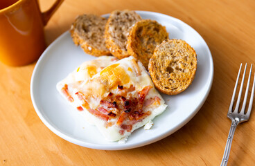 Square pieces of egg and bacon omelet casserole. Meal is served with rye bread and earthenware cup of tea..