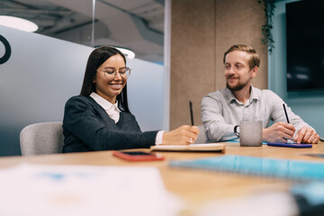 Smiling young woman writing notes in collaborative meeting while male colleague listens, showcasing digital teamwork culture and creative thinking in tech-first professional space