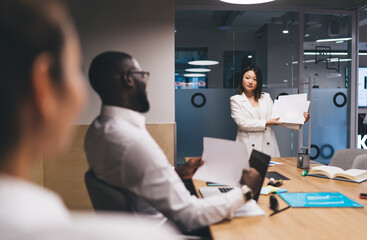 Focused Asian businesswoman in blazer leading team presentation with printouts, emphasizing business communication and data sharing in a collaborative, tech-integrated office setting