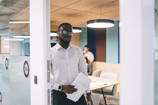 African American businessman enters conference room holding documents and tablet, preparing for technology-driven business presentation in collaborative workspace