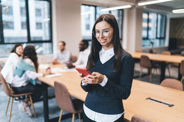 Professional woman in glasses using red smartphone, smiling warmly in bright coworking office, engaging in digital communication during workday with team in background