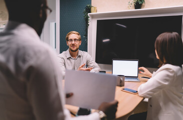 Man explaining project details during team meeting in startup office, standing with report in hand,...