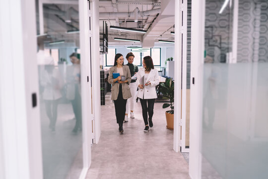 Smiling female coworkers walking together through modern office hallway, carrying notebooks and coffee, engaged in relaxed business conversation in friendly tech company environment