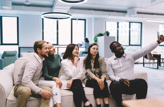 Young diverse professionals smiling while taking group selfie on office couch, creating lasting memory of team spirit, modern work culture and connection in casual workspace
