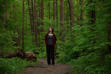 Obraz premium Beauty face young woman with serious thinking look standing on the path among green pine trees in the forest. Girl walking and looking up in woods Emotion closeup full length portrait