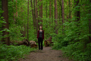 Obraz premium Beauty face young woman with serious thinking look standing on the path among green pine trees in the forest. Girl walking and looking up in woods Emotion closeup full length portrait