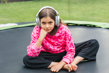 Girl 8-9 years with serious expression in pink sweatshirt and headphones sitting cross-legged on trampoline outdoors, concept of childhood, theme of emotions, music, personality