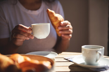 A close-up view of a woman holding a coffee cup and croissant, enjoying a peaceful morning breakfast under warm sunlight. Coffee and relaxation in a serene atmosphere.