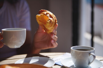 Close-up of a person enjoying a coffee and croissant outdoors. Warm sunlight creates a cozy...