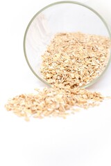 A clear glass tipped over, with raw rolled oats spilling onto a white surface. The clean minimalistic composition highlights the texture and natural golden color of the oats, commonly used for healthy