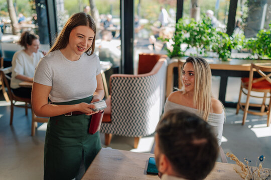 Smiling waitress holding pos terminal accepting payment from customers in restaurant