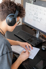 Teenager studying math online with headphones, taking notes at desk. Distance learning, school...