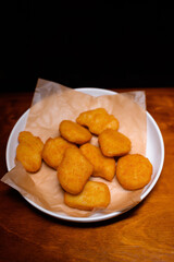 Golden crispy chicken nuggets served on parchment paper in a white plate on a wooden table. Tasty fast food snack, perfect for parties or quick meals
