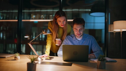 Smiling partners looking laptop at dark office closeup. Happy managers rejoicing