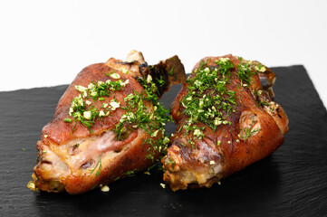 Two roasted pork knuckles with crispy brown crust, generously topped with fresh chopped dill and garlic, served on a stylish black slate plate over a clean white background
