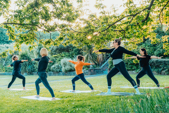Female yoga class in park. Group of diverse women doing stretching pose exercising together with instructor on green grass lawn. Sport activity for health, wellbeing, mental health. Selective focus