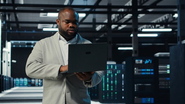 Programmer in data center using laptop, testing equipment failover scenarios. Server farm IT professional uses software on notebook, ensuring data accessibility, camera B