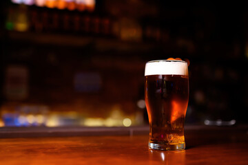 A glass of dark beer with a creamy foam stands on a wooden table in the center of the frame. The bar area is bathed in soft red light, creating a cozy and intimate atmosphere