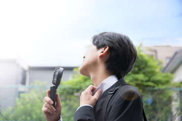 暑い中汗をかく営業する大変なビジネスマン　ハンディファンで涼むイメージ　夏の外回りは熱中症に注意