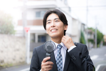 暑い中汗をかく営業する大変なビジネスマン　ハンディファンで涼むイメージ　夏の外回りは熱中症に注意