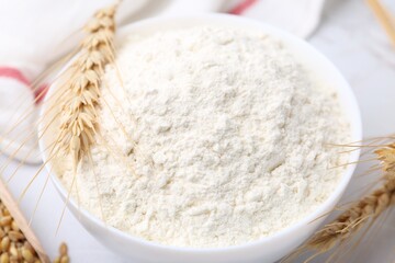 Wheat flour in bowl, grains and spikes on table, closeup