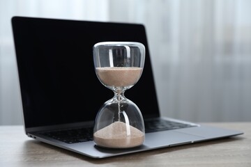 Deadline. Hourglass with flowing sand and laptop on wooden table indoors, closeup