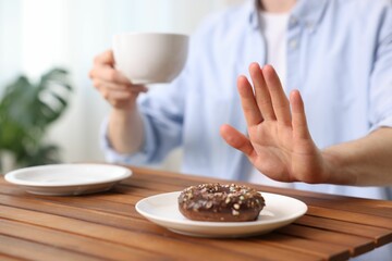Gluten free diet. Man with cup of drink refusing from donut at wooden table indoors, closeup