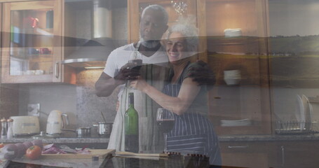 Toasting couple wearing aprons cooking in home kitchen, with wine glasses, cutting board, knife