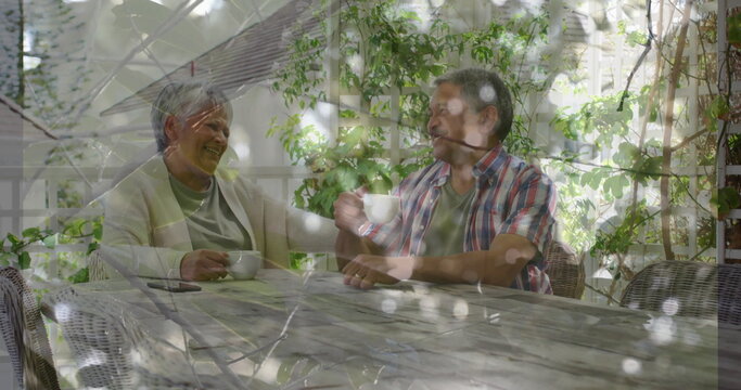 Sitting senior couple holding white coffee cups and checking phone on garden patio under pergola