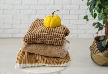 Stool with stack of different stylish sweaters, pumpkin and houseplant near light brick wall in room