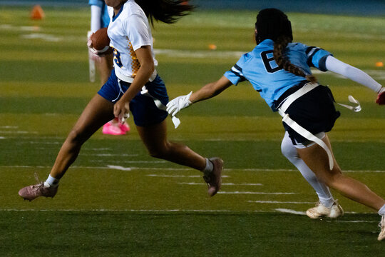 Close-up of intense flag football play, girl sprinting with ball as opponent lunges to grab flag, dynamic youth sports action, girls in motion, athletic energy, competition and pursuit on turf.