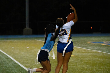 Female flag football receiver leaps for catch under pressure, with defender closing in during intense nighttime match. Athleticism, focus, and sportsmanship captured on artificial turf