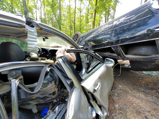 Two vehicles are severely damaged in wooded area, with one car flipped over debris scattered on ground.