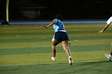 Girls flag football player in blue jersey makes a defensive move under stadium lights, focused positioning and readiness during night game, youth athleticism, school competition, action capture.