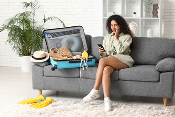 Beautiful young happy woman with mobile phone unpacking summer clothes from suitcase at home