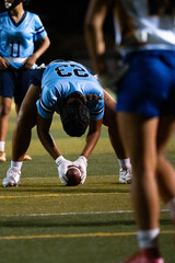 Girls flag football center prepares to snap the ball at night game, crouched in position with focus and intent, youth athletes in action under lights, sports readiness, school competition scene.