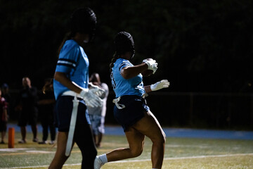 Girls flag football player with braided hair carries ball under stadium lights, preparing to celebrate a touchdown, nighttime school sports action, determination, energy, teamwork and success.