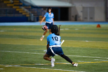 Girls flag football receiver in blue jersey crouches to catch pass during night game, intense sports action on turf, youth athletic focus, school competition, teamwork and coordination captured.