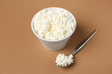 Bowl and spoon with fresh cottage cheese on brown background