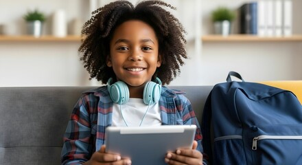 Young boy smiles holding tablet with headphones and backpack