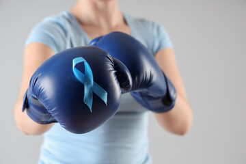 Young woman in boxing gloves with blue ribbon on light background, closeup. Cancer awareness concept