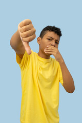 Vertical image of a dark skinned Latino teenager wearing a yellow shirt with a disappointed face making a thumbs down gesture on a light blue background with copyspace