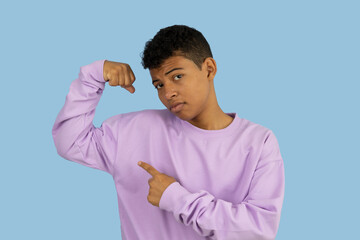 Young brown skinned Latino man making a gesture of strength with his hands, he is wearing a lilac sweatshirt and points his index finger at the muscle on a light blue background