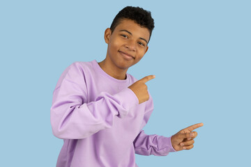 Cheerful dark skinned Latino teenager wearing a lilac sweatshirt pointing with his index fingers at a copyspace on a light blue background, young man indicating an advertising space
