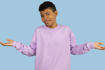 Dark skinned Latino teenager wearing a purple sweatshirt spreading his hands with a questioning expression on a light blue background, confused young man concept