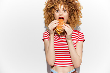 Young woman with curly hair excitedly biting into a delicious burger, wearing a striped crop top...