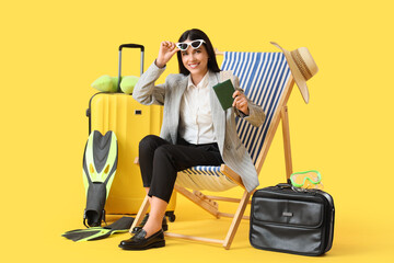 Young businesswoman with passport in deck chair and beach accessories on yellow background. Summer...