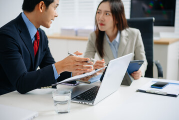 Two Asian business professionals in formal suits discussing a project at modern office desk with laptop and documents.